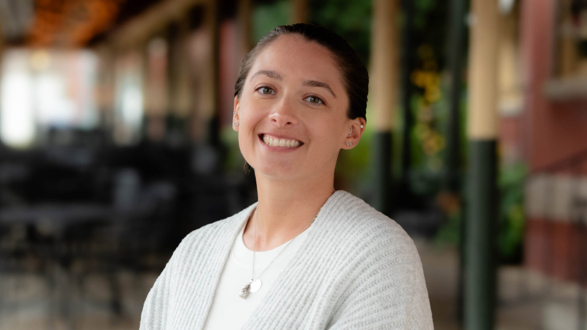 A professional photo of Shannon Maria, a woman with light skin and dark hair wearing a gray sweater. She is smiling and standing in front of a brick building.